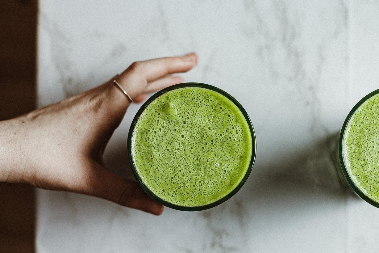 Two green smoothies in glass cups on a marble surface with a hand holding one of them.